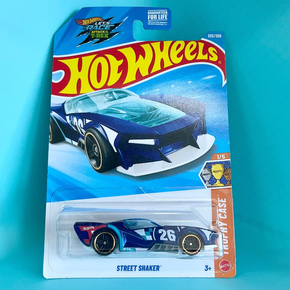 A Hot Wheels Street Shaker toy car in its original blister card packaging, set against a solid turquoise background. The card features the classic red and yellow Hot Wheels logo and depicts the blue and white race car with the number 26.