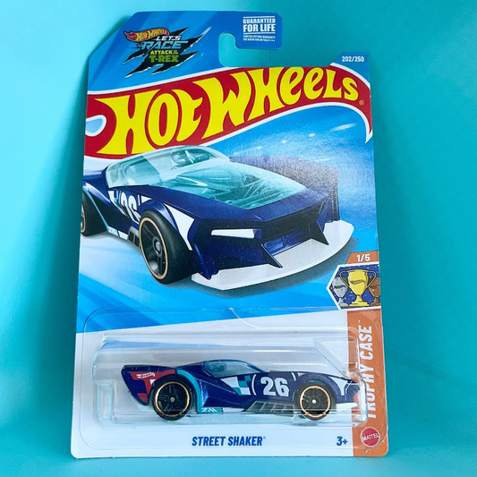 A Hot Wheels Street Shaker toy car in its original blister card packaging, set against a solid turquoise background. The card features the classic red and yellow Hot Wheels logo and depicts the blue and white race car with the number 26.